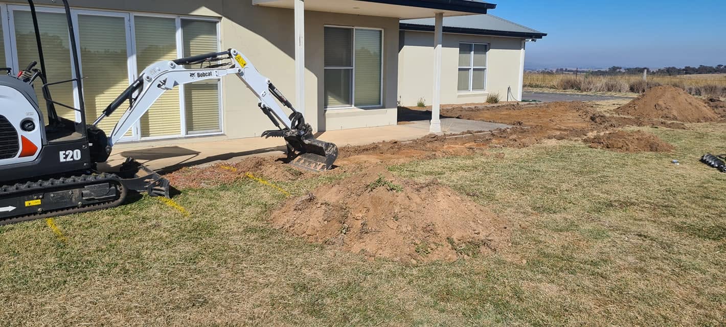 Bobcat excavator trenching for drainage alongside a rural home